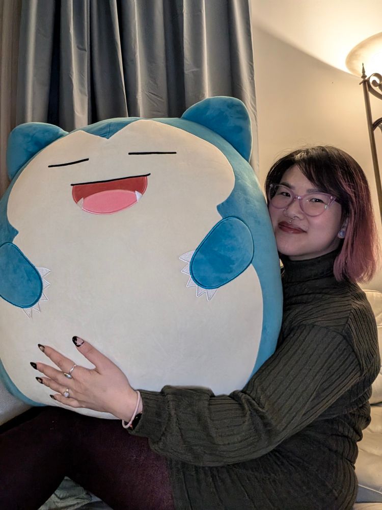 A woman with short pink and black hair and purple glasses has a slight smile on her face. She is holding a giant plushie of Snorlax. The plushie rests on her body. She is lounging on top of a white couch.