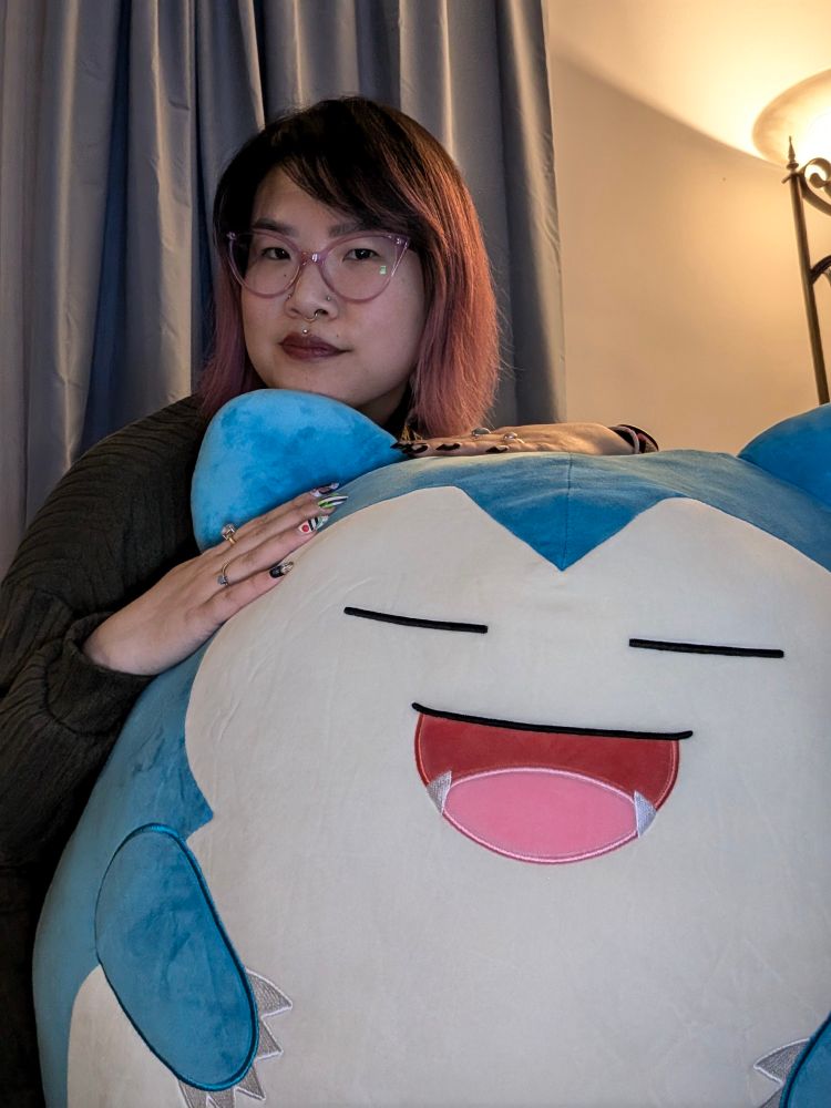 A woman with short pink and black hair and purple glasses has a hint of a smile on her face. She is resting her head and hands on top of a giant plushie of Snorlax. She is on her knees to appear taller than the plushie. 