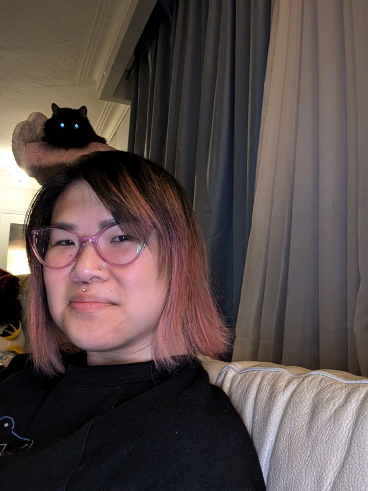 Photo of me (with short dusty pink and black hair with translucent purple cateye glasses) with a slight smile and sitting on a white leather couch. Behind me is my black cat perched on her cat tree, and with the camera's flash, her only features you can see are her two glowing blue eyes.