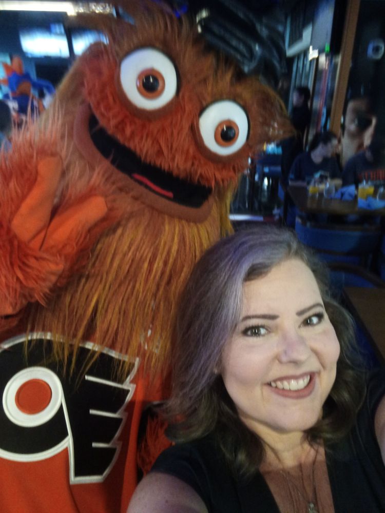 A selfie of Manda, a while re woman with purple hair, with Gritty, the Philadelphia Flyers mascot - who is an orange, shaggy, muppet-like creature 