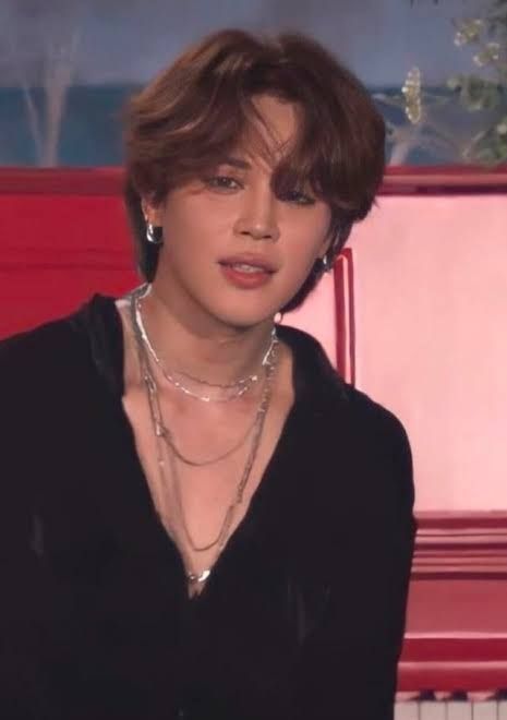 Jimin in low cut black shirt and lots of chains sitting in front of red piano