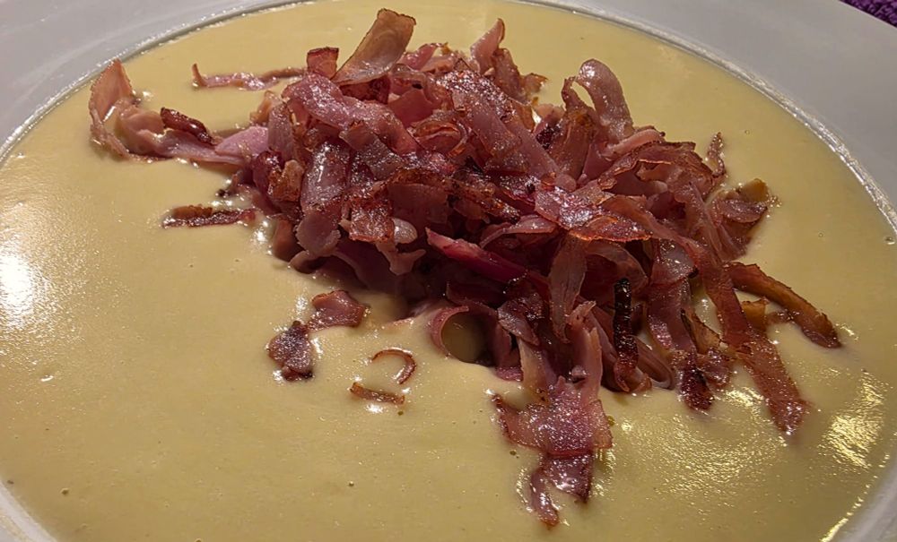 Nahaufnahme eines Tellers mit heller Cremesuppe, darin liegen angebratene Streifen von Schinken.