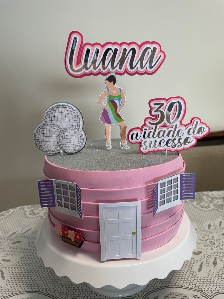 Bolo fake redondo cor de rosa, imitando a casinha do filme “De repente 30” com duas janelas e uma porta branca ao centro, e um vaso de flores abaixo da janela esquerda, e com topo de bolo (“Luana” ao centro, com uma figura da Jenna na frente, a esquerda balões metálicos e a direita escrito “30 a idade do sucesso”) 