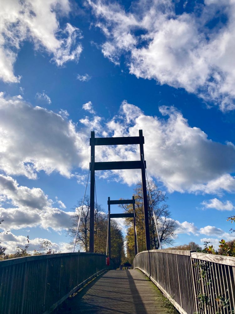 A posted footbridge in the sunshine surrounded by blue skies and scudding soft white clouds, tinged with grey 