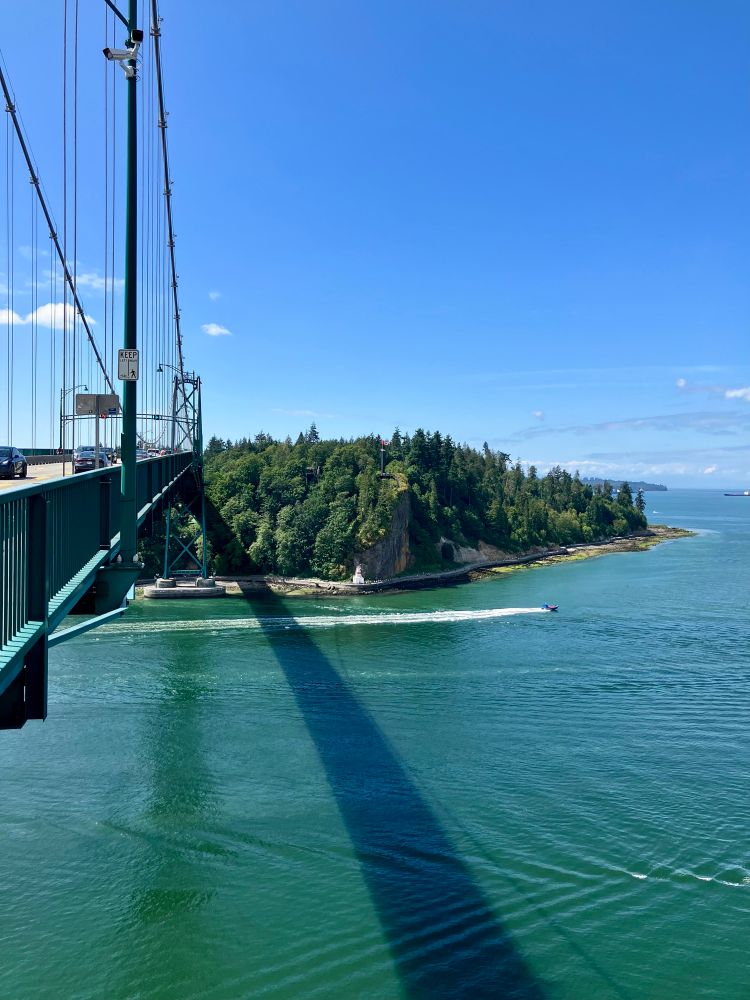 Stanley Park from mid-span on the Luon’s Gate bridge. The green metal structure of the bridge casts a shadow on the green water of the First Narrows below. 