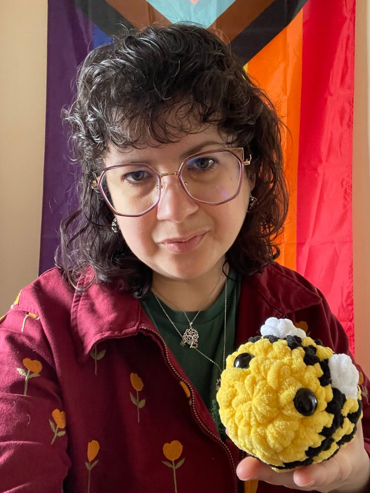 El, a white passing person with midlength dark brown hair is taking a selfie in front a progress pride flag. They are wearing purple framed glasses and are holding a cute squishy bee. 