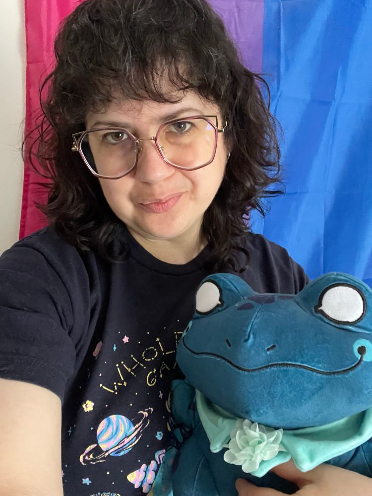 El, a white passing person with midlength dark brown hair is taking a selfie in front of a bi pride flag. They are wearing purple framed glasses and are holding a Atul plush from Spiritfarer.