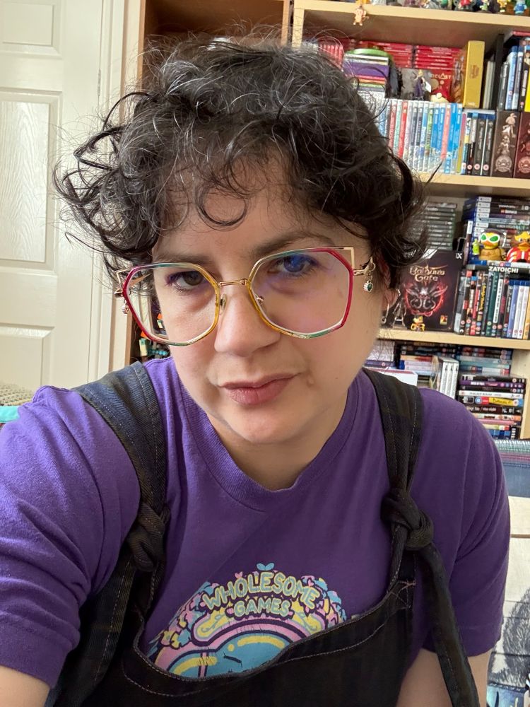 El, a white passing person with midlength dark brown wavy hair tied up is taking a selfie in front of shelves that have games and Lego figures on them. They are wearing rainbow framed glasses and a purple logo T-shirt.