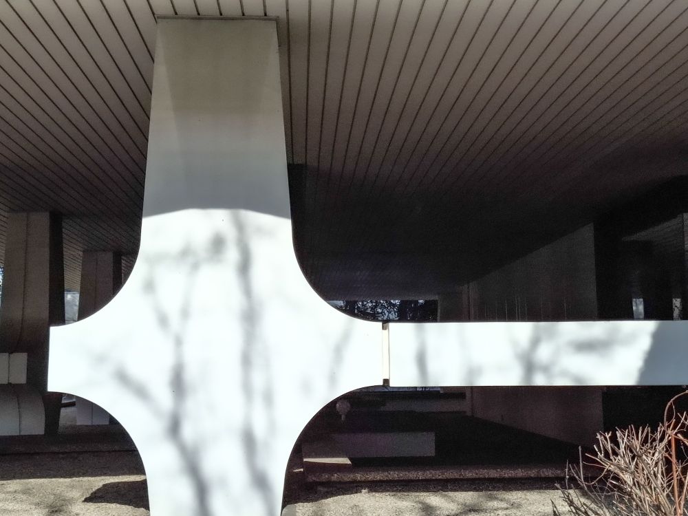 Élément architectonique. Poteau en forme de croix sous un immeuble de standing des années 70. La sous face de la dalle est en lames métalliques blanches. Le poteau est en béton blanc sculptural.