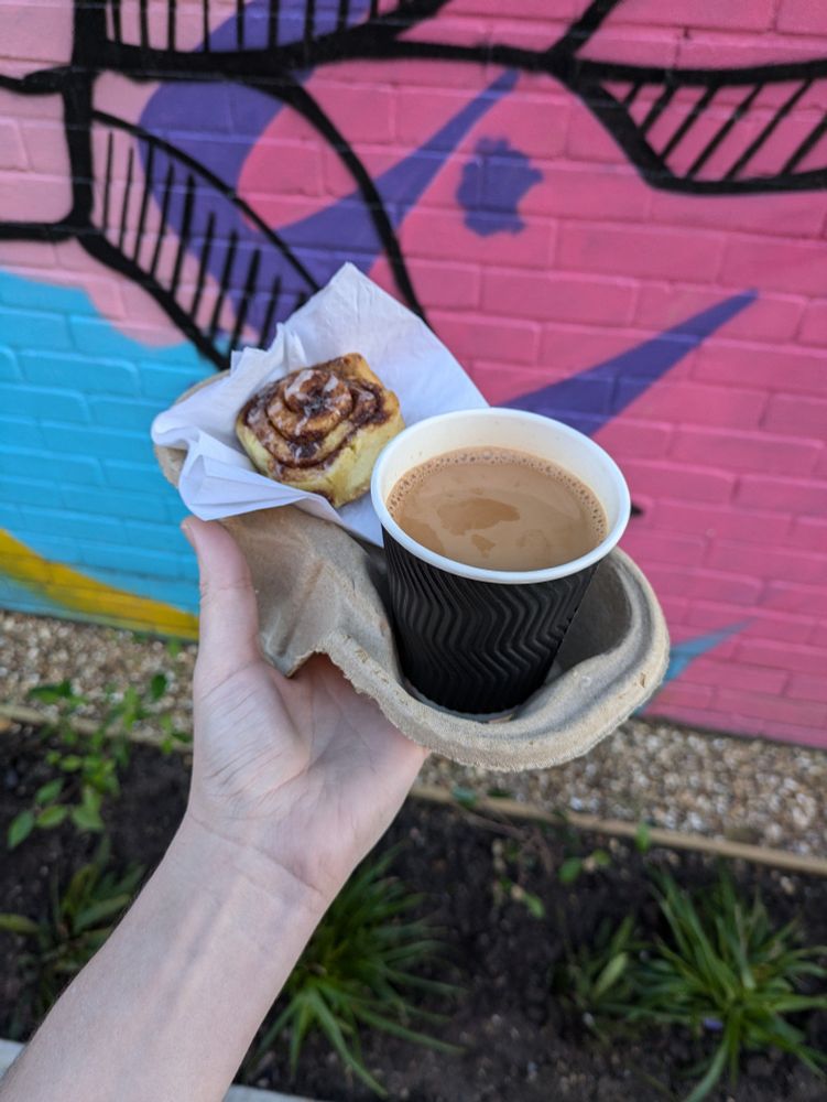 My hand holding up a cinnamon roll and a Karak chai outside. It the background is a graffitied wall with some plants. 