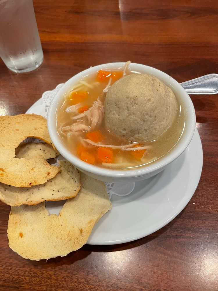 Matzo ball soup in a white bowl on a white plate on a white doily with a side of bagel chips. The matzo ball is large and fluffy and sitting in broth with chunks of carrots, pieces of chicken, and a few noodles. 