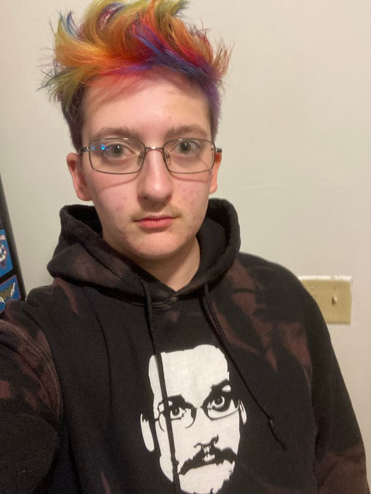 Lys is wearing a Pizza John hoodie and are looking at the camera with a blank stare. They have a rainbow Mohawk and a brown mustache and are wearing glasses similar to John’s.  