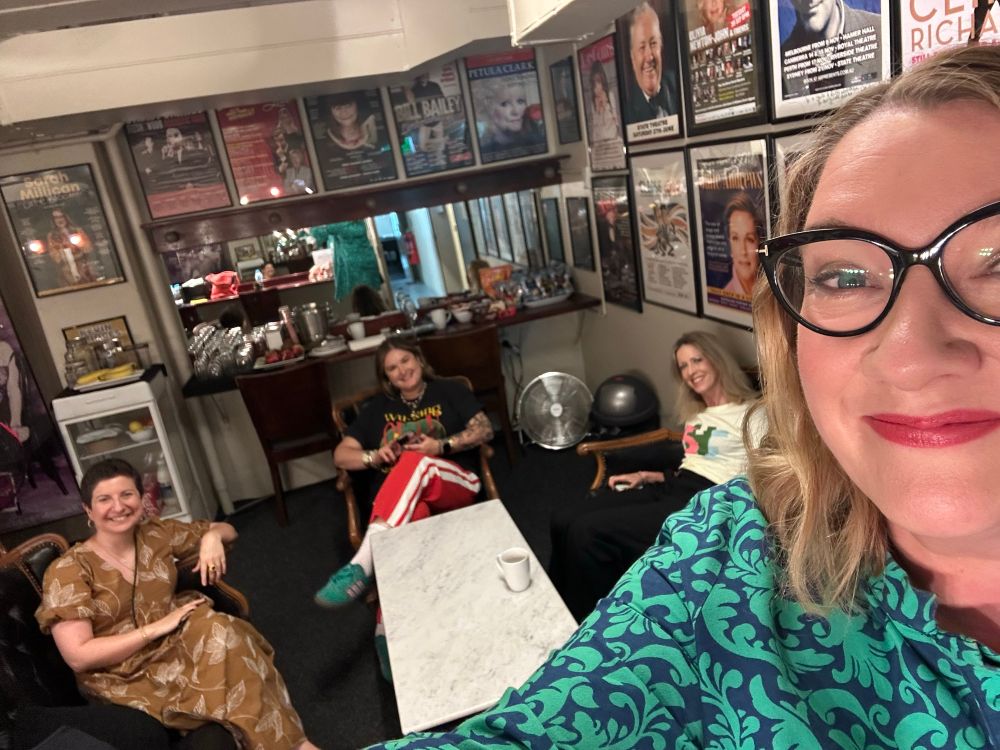 It’s a glimpse of the backstage area. The dressing room walls are adorned with posters of people who’ve been there before. There’s a coffee table and three excellent women sitting in comfy chairs around it. I’m in the foreground in a blue and turquoise Zuri dress with pink lipstick and wavy (day 2) hair. 