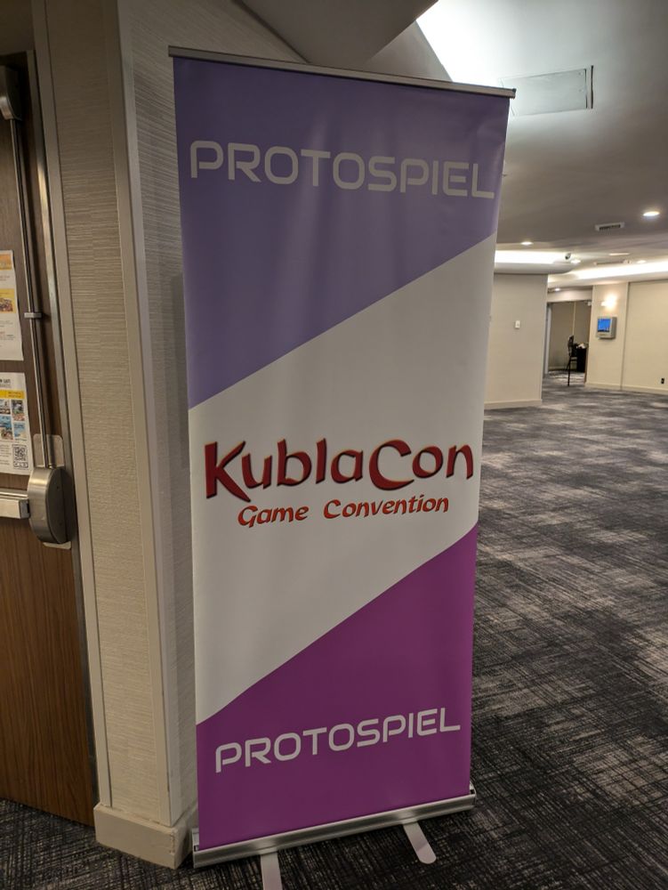 A Protospiel banner at KublaCon. It's vertically oriented, with a lavender, white and purple stripes.