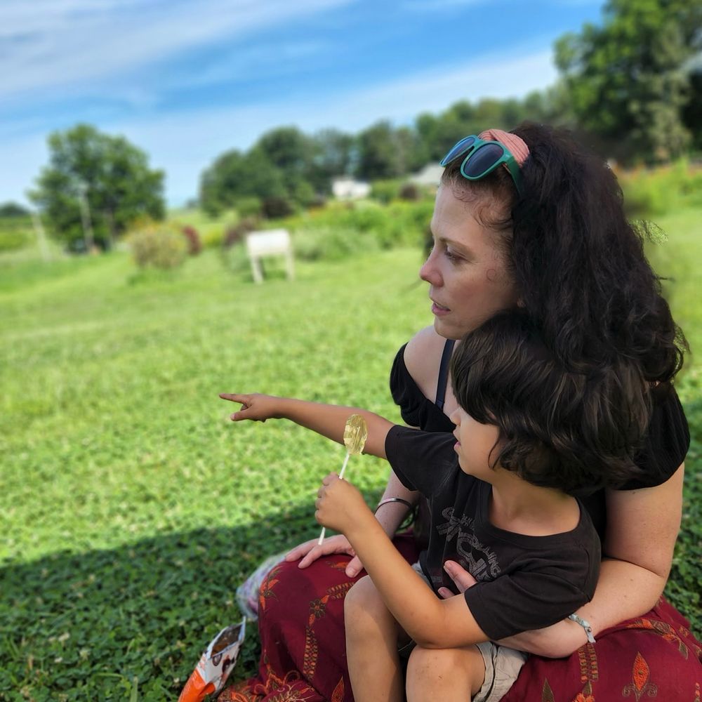 A profile shot of my 5yo son sitting in my lap in a green field. In one hand he has a lollipop, with the other he’s pointing off into the distance and I’m looking off in that direction. 