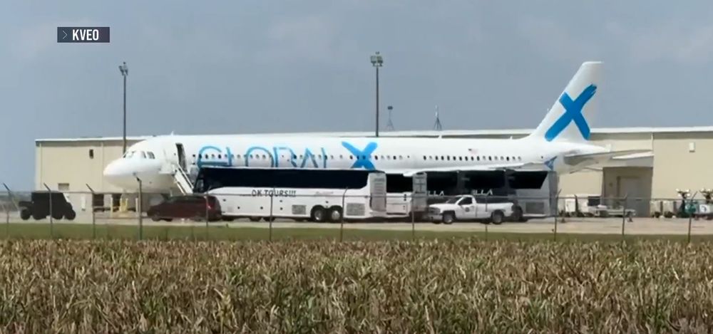 A Global X A321 with a bunch of charter buses parked around it. There's an airport hangar in the background and dried brush in the foreground.