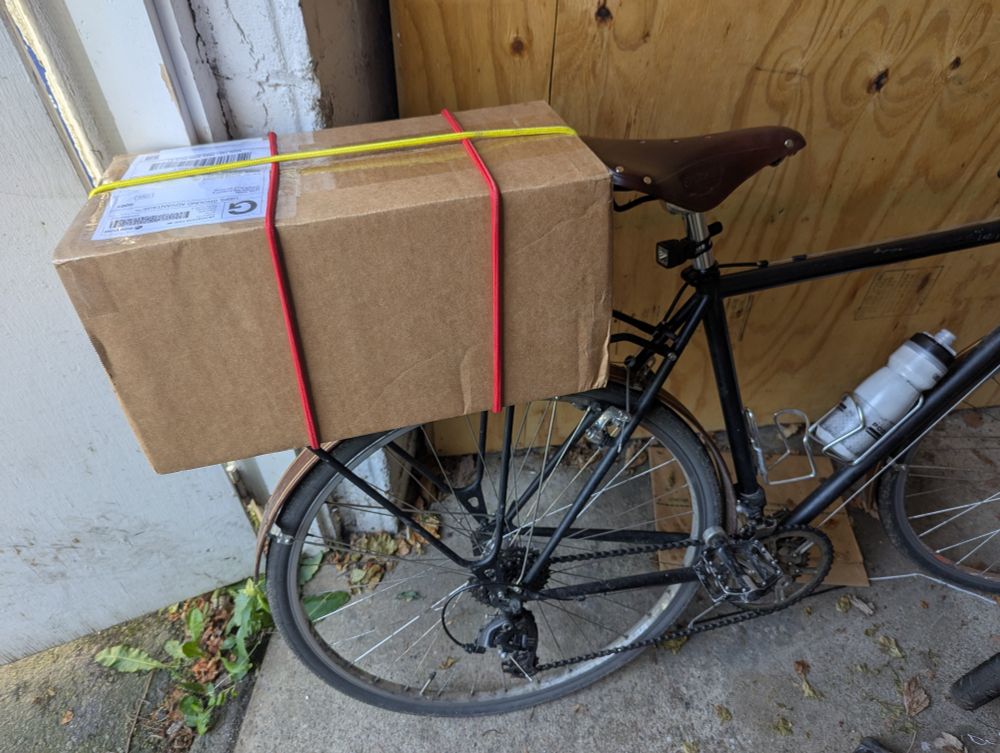 Cardboard box bungeed to the rear rack of a black bicycle leaning against a garage wall. The bungee cords are red and yellow.