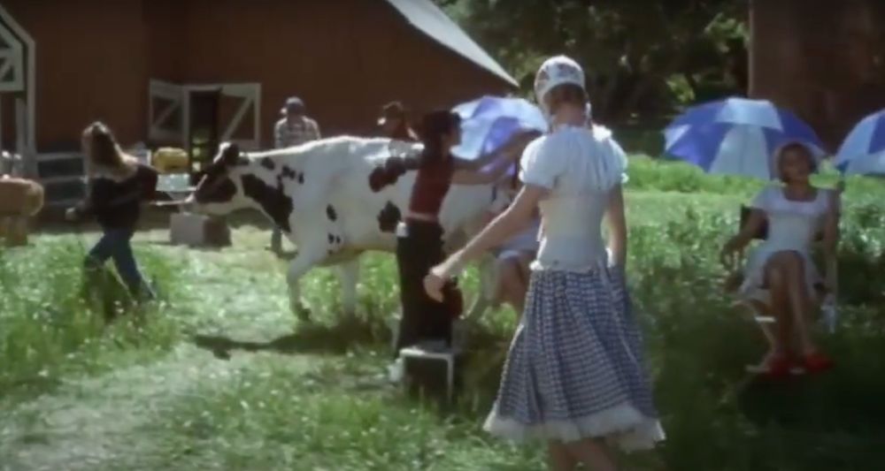 A screenshot from The Truth About Cats and Dogs, showing Uma Thurman in a milkmaid outfit in front of a barn, several other people, and a white cow with black spots. 