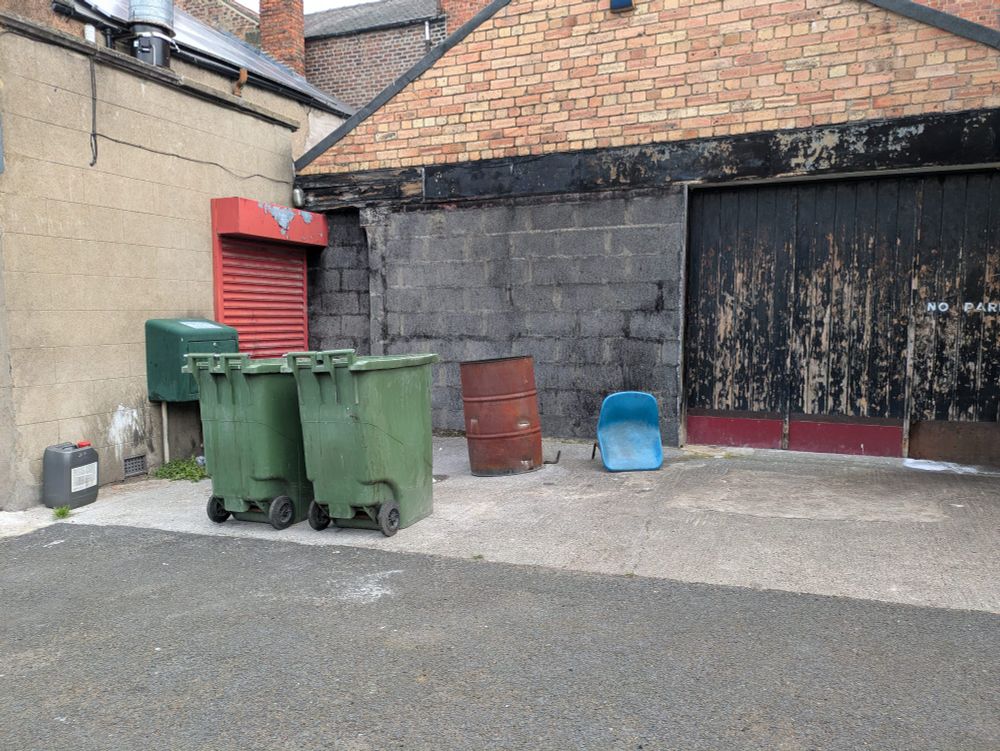 This is a corner of Hexham, Northumberland that interested me this morning. There is a red shutter behind some green wheelie bins neatly arranged behind a grey tarmac side road and in front of a single storey building.