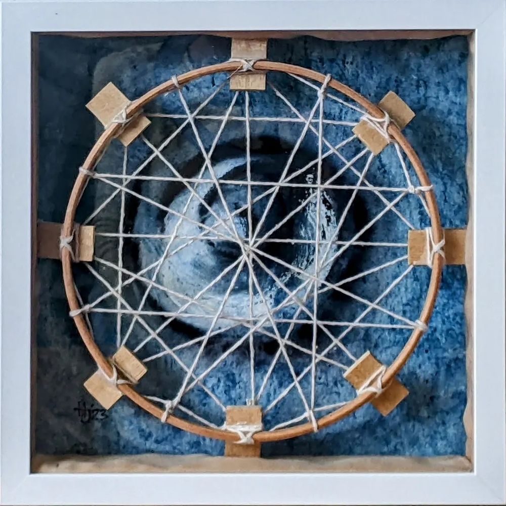 This is a mixed media piece of artwork that was inspired by looking up at the top of the roof of the Yurt Café at Limehouse, London from the inside. You can see the sky through the opening of the tent and the perspex that covers the opening rattles when the wind blows.

The colours on the background are mainly blue and black and painted on handmade Indian paper and the tent is described using an embroidery hoop with string woven through it to create a geometric pattern. I believe I made it in 2023.