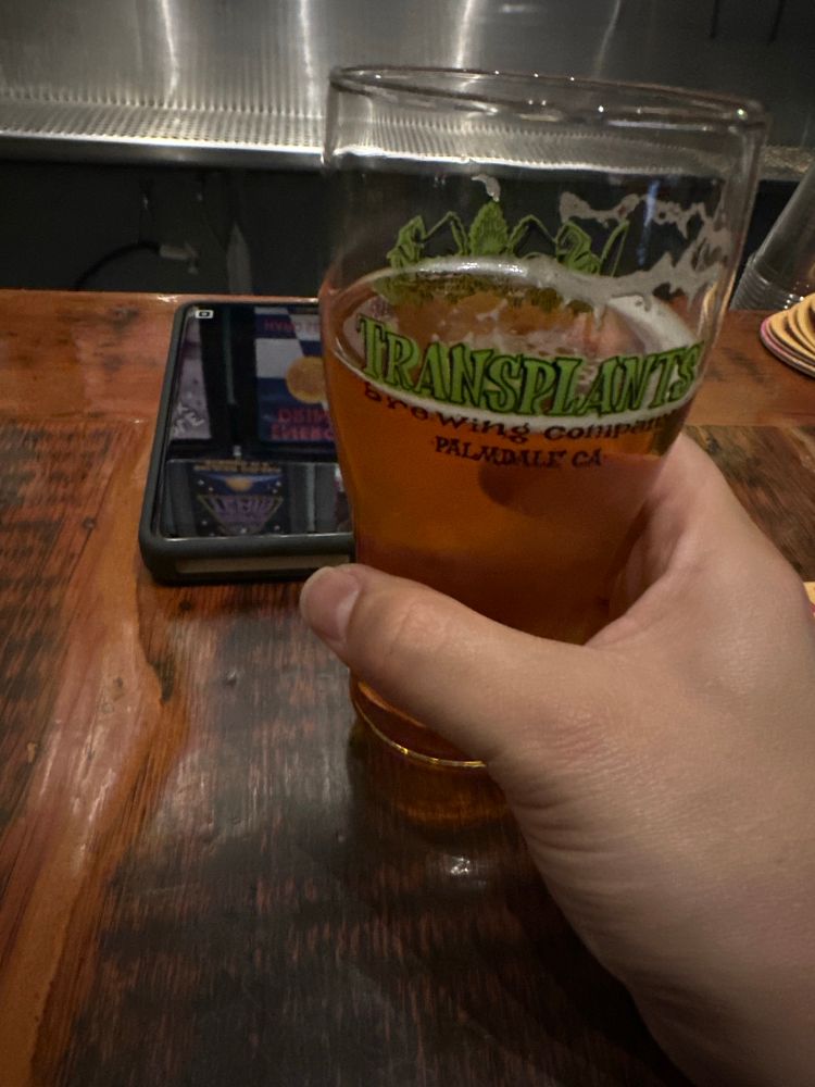 My hand holding a glass of beer, filled with Transplants Brewing’s Mas Guapo IPA with Guava (7.5%)