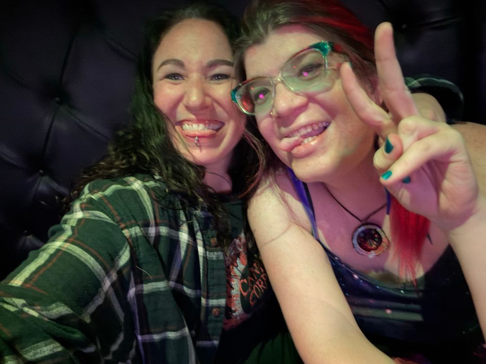 Two beautiful women with their tongues out. The one on the left has a great toothy smile, curly dark hair, cannibal corpse shirt under a green plaid flannel button down shirt. She also has her tongue pierced and her arm around the woman on the right who has rainbow glasses, spaghetti strap dress, blue nail polish. The girl on the right also has a rainbow slice of pine cone necklace and a nose ring. 