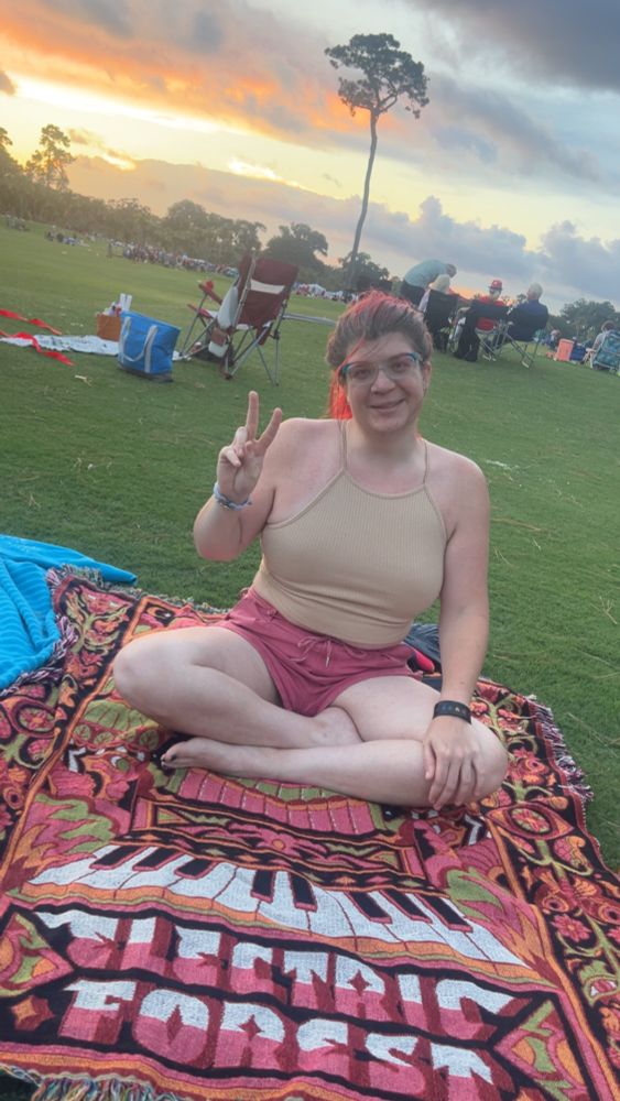 This user on a pink Electric Forest Blanket. On a driving range with a lone tree in the background with the sunset that has deep oranges and golden yellows. Ok so the color scheme of this photo is the Lesbian Pride Flag. 