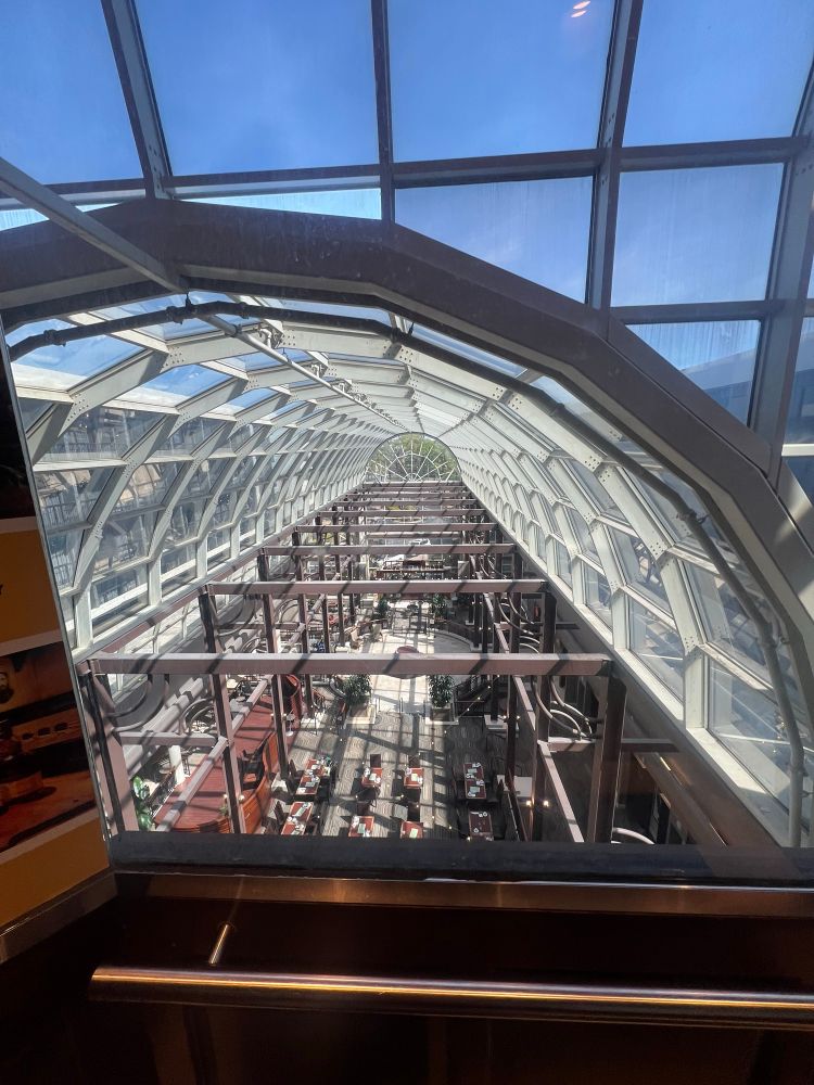 Hotel atrium viewed from elevator on 6th floor with a glass arched roof and a semicircle of glass panels sliced like a pie on the other end. Metal structure is visible with atleast 9 beams at regular intervals starting at the bottom, flat part of the semicircle arched roof. Dining area with set tables visible below and a bar at the end. Blue sky is visible thru the roof which is entirely made of metal structure and glass panels.