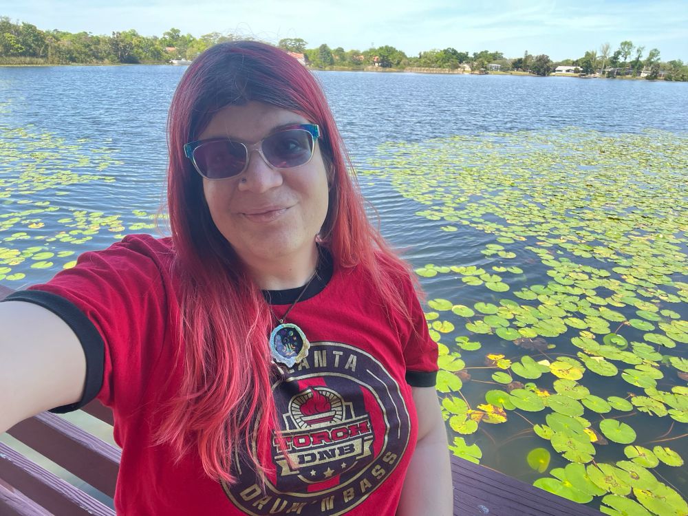 Woman with long red hair and rainbow sunglasses in a red, black and gold shirt that says Atlanta Torch DNB Drum n Bass, in a circular logo on n the middle of her shirt. A lake is in the background with tons of lillypads on the water to either side behind her. The sky is blue and there is a shoreline with houses and large trees on the far side of the lake in the background. 