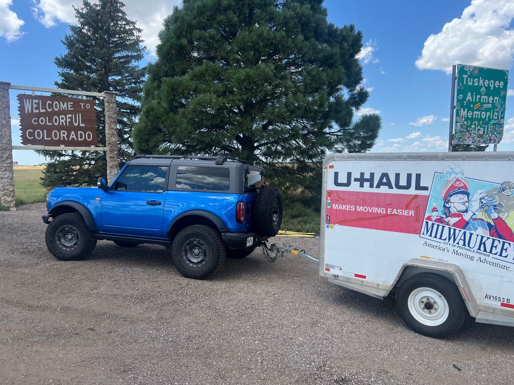 “Welcome to Colorful Colorado” sign with a blue 2 Door Bronco towing a U-Haul trailer. Blue skys and some trees in the background.