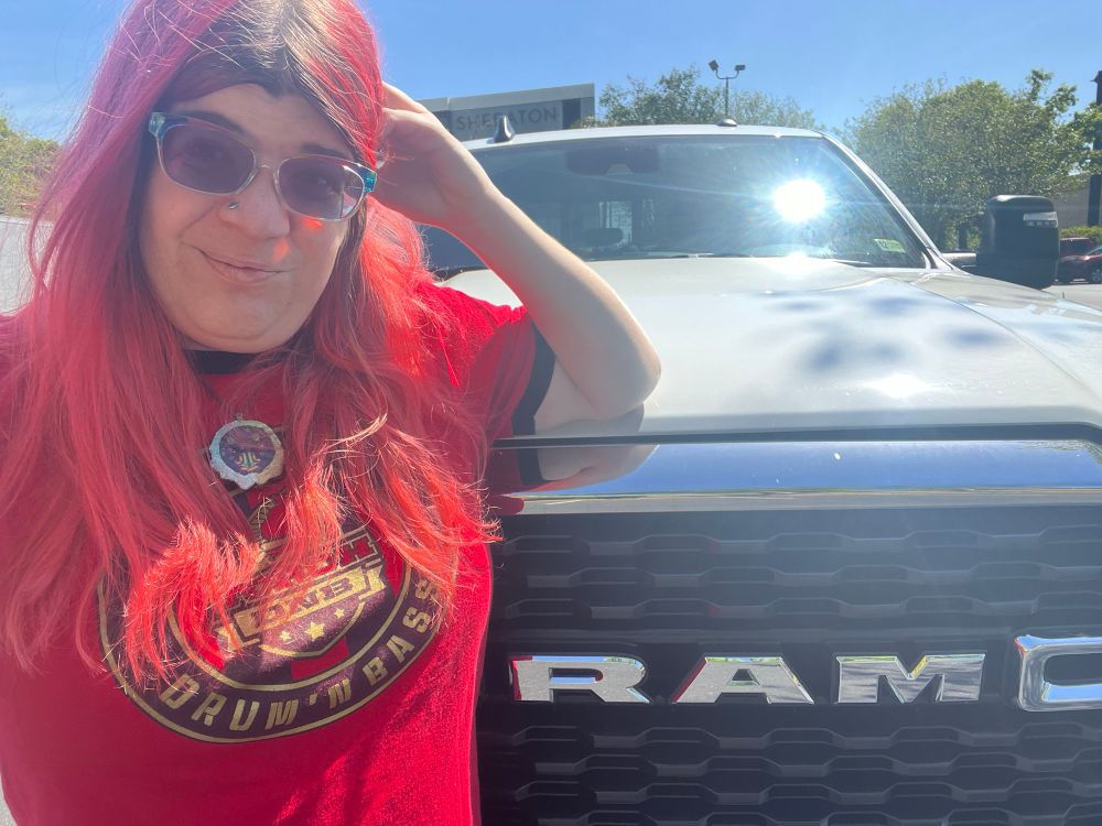 Woman with long red dyed hair and rainbow sunglasses leaning her arm on the hood of a truck that comes up to shoulder height. RAM is visible on the grill of the white truck. The woman’s red shirt says Drum n Bass