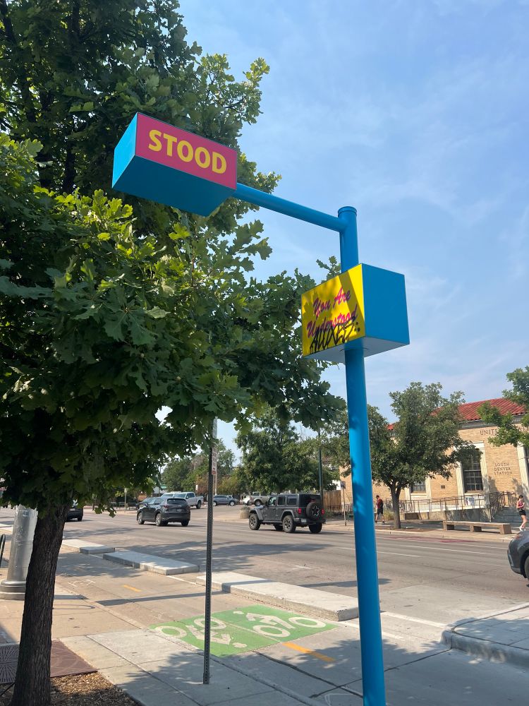 Blue pole on the sidewalk next to a tree that says “you are understood” with a blue box hanging over the sidewalk that says “Stood” blue skies