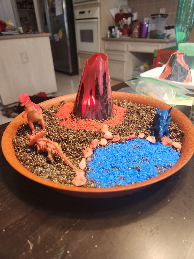 Dinosaur display with a small volcano in the center and a blue pond on the bottom right. The ground is currently dirt with plants expected to grow in.