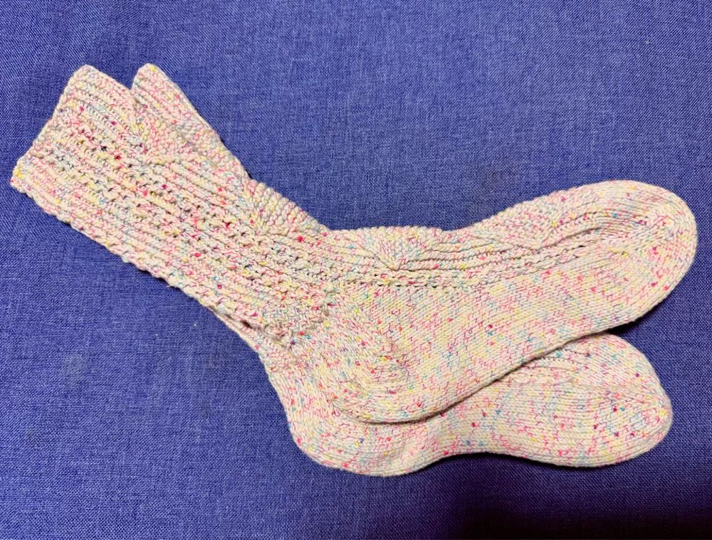 青い座布団に手編みの靴下を左右重ねて置いた写真。靴下は白地に虹色が混ざったような糸で大きなダイヤマークの模様編みになっている。