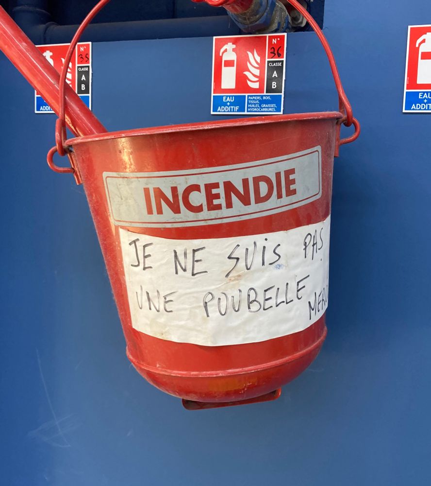 A fire bucket in a French supermarket with a handwritten note on it reading JE NE SUIS PAS UNE POUBELLE