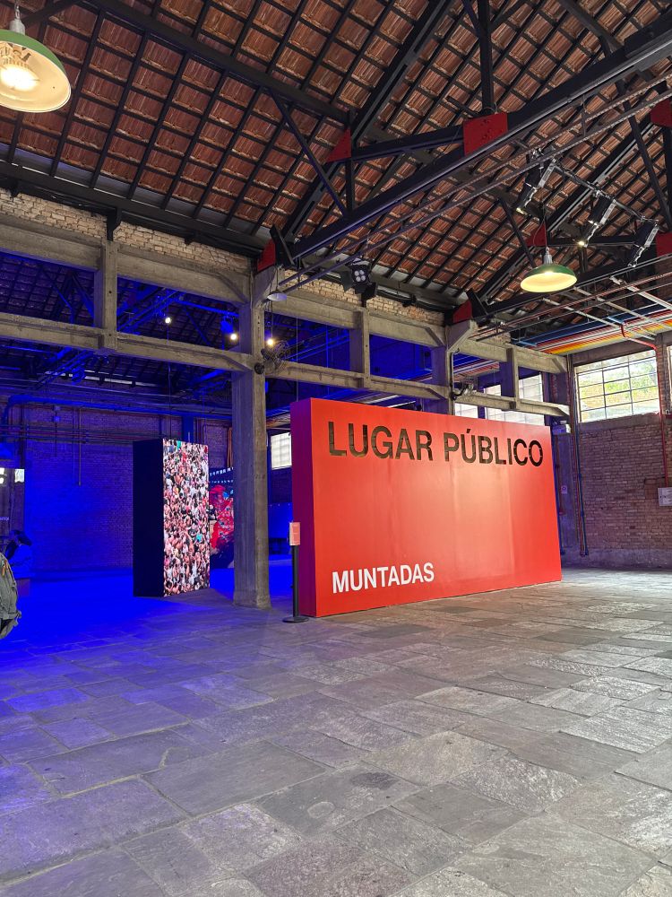 Foto de dentro de um galpão iluminado com luzes amarelas e azuis e na frente um muro vermelho com texto “Lugar público” 