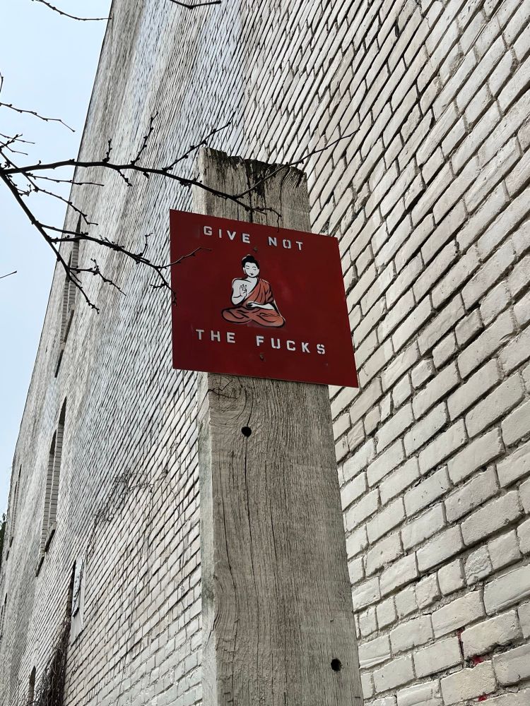 A red piece of metal with a spray-stenciled image of a person in a draping robe seated with their legs crossed, one hand raised in front of their chest. Above and below are the spray-stenciled words “GIVE NOT THE FUCKS”