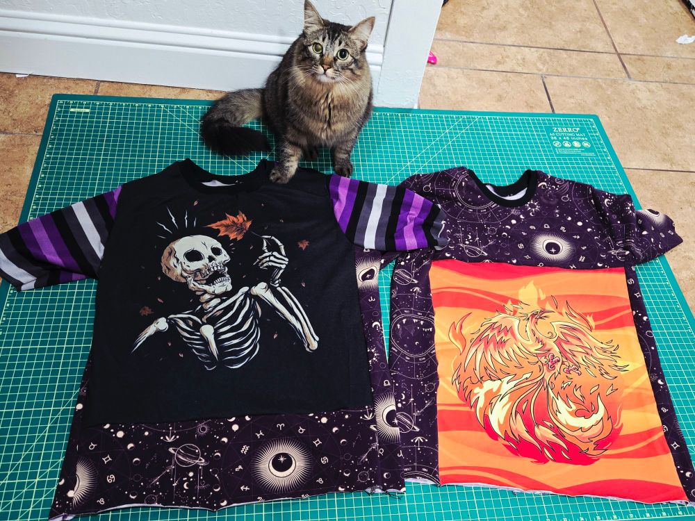 A fluffy tabby cat posing on a cutting board next to a shirt with a skeleton holding up a leaf, purple and black striped sleeves, and astrological print at the bottom. On the right is a shirt with a big phoenix panel and astrological print. 
