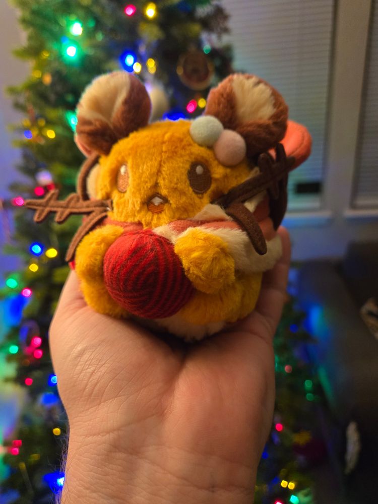 Picture of my hand holding a holiday Dedenne plush. He's holding a ball of yarn and wearing a holiday coat. 