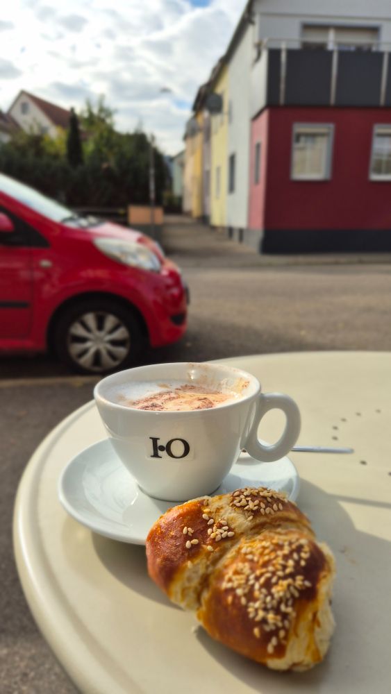 Caféhaustisch im Freien an Straße mit Tasse Cappuccino und Sesamcroissant