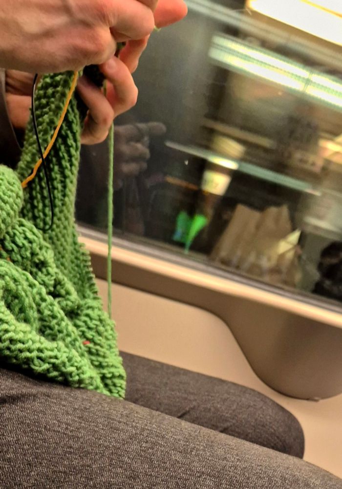 Männeroberschenkel auf Stadtbahnsitz, darüber seine Hände an grünem Strickzeug