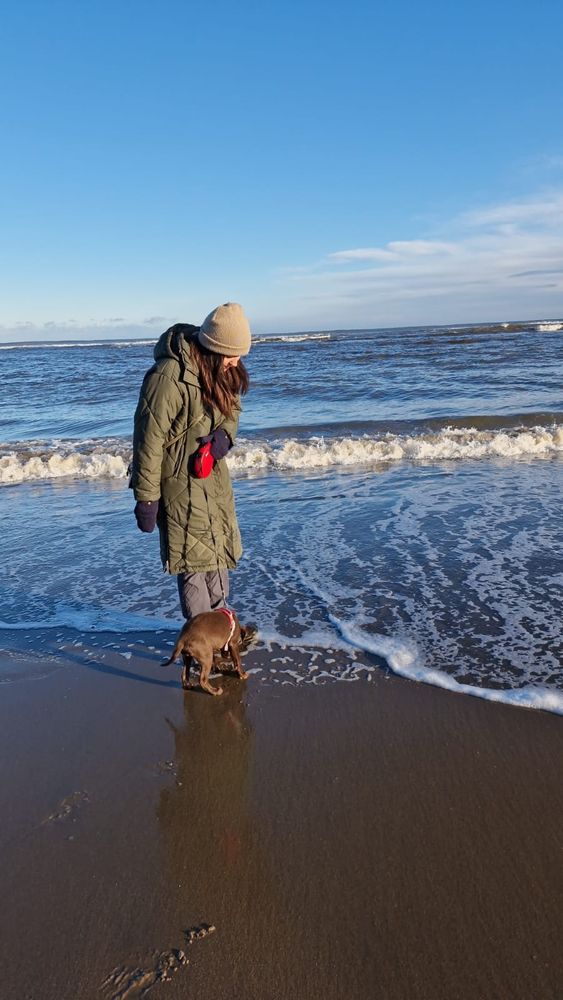 Alice (a white woman with dark hair, wrapped up in a duvet-coat, gloves & hat) on the beach with a 3mo old chocolate labrador. The sea is about to roll over their feet.
