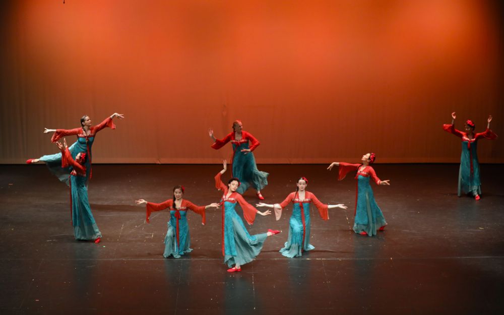 Women in teal and rust dresses dance on a stage with an orange background.