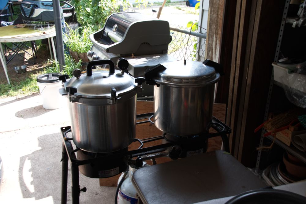 Two pressure canners of different manufacture stand side by side on a camp stove in front of an open garage.  A grill is outside behind them.