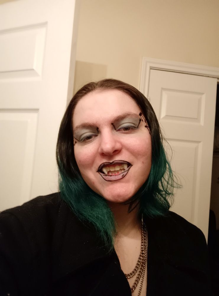 An androgynous young adult in a black captain's coat. They are wearing small vampire fangs.
Hair is styled short to the shoulders, brown tipped with teal. They are wearing light blue eyeshadow, have computer lines drawn on either side of the face at the temples, and black outlining on their lips.