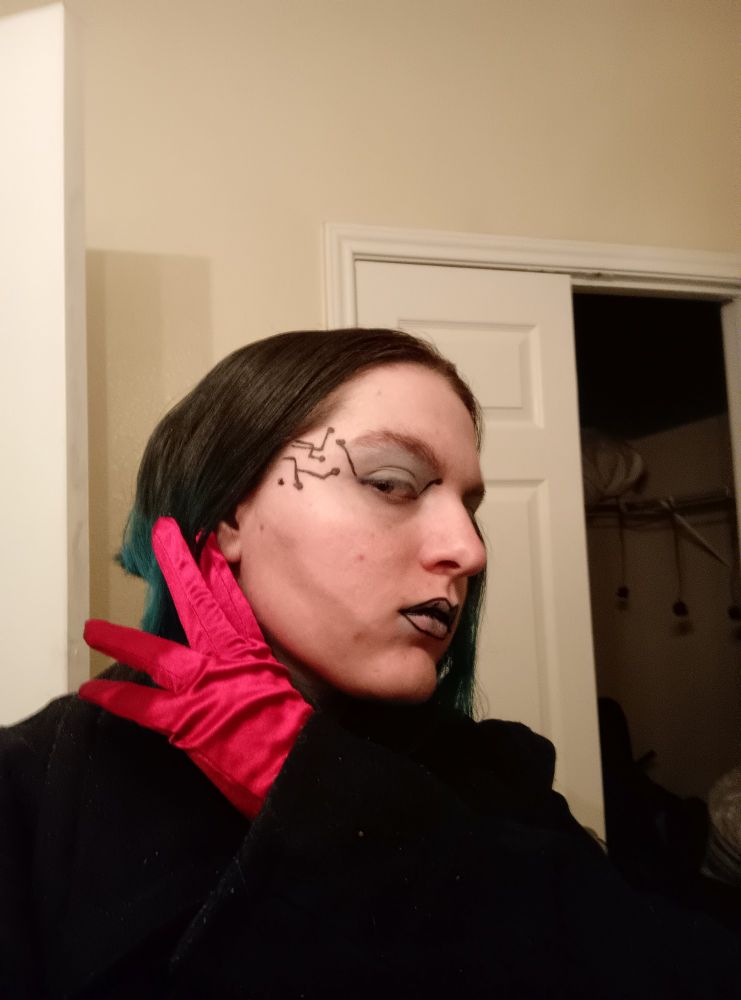 An androgynous young adult in a black captain's coat, wearing a single red glove.
Hair is styled short to the shoulders, brown tipped with teal. They are wearing light blue eyeshadow, have computer lines drawn on either side of the face at the temples, and black outlining on their lips.
Their head is tilted to one side to show the computer line details.