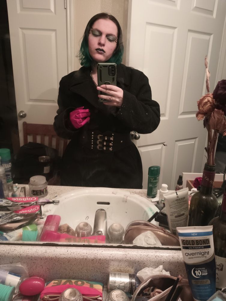 An androgynous young adult in a black captain's coat with a silver buckle belt, wearing a single red glove.
Hair is styled short to the shoulders, brown tipped with teal. They are wearing light blue eyeshadow, have computer lines drawn on either side of the face at the temples, and black outlining on their lips.