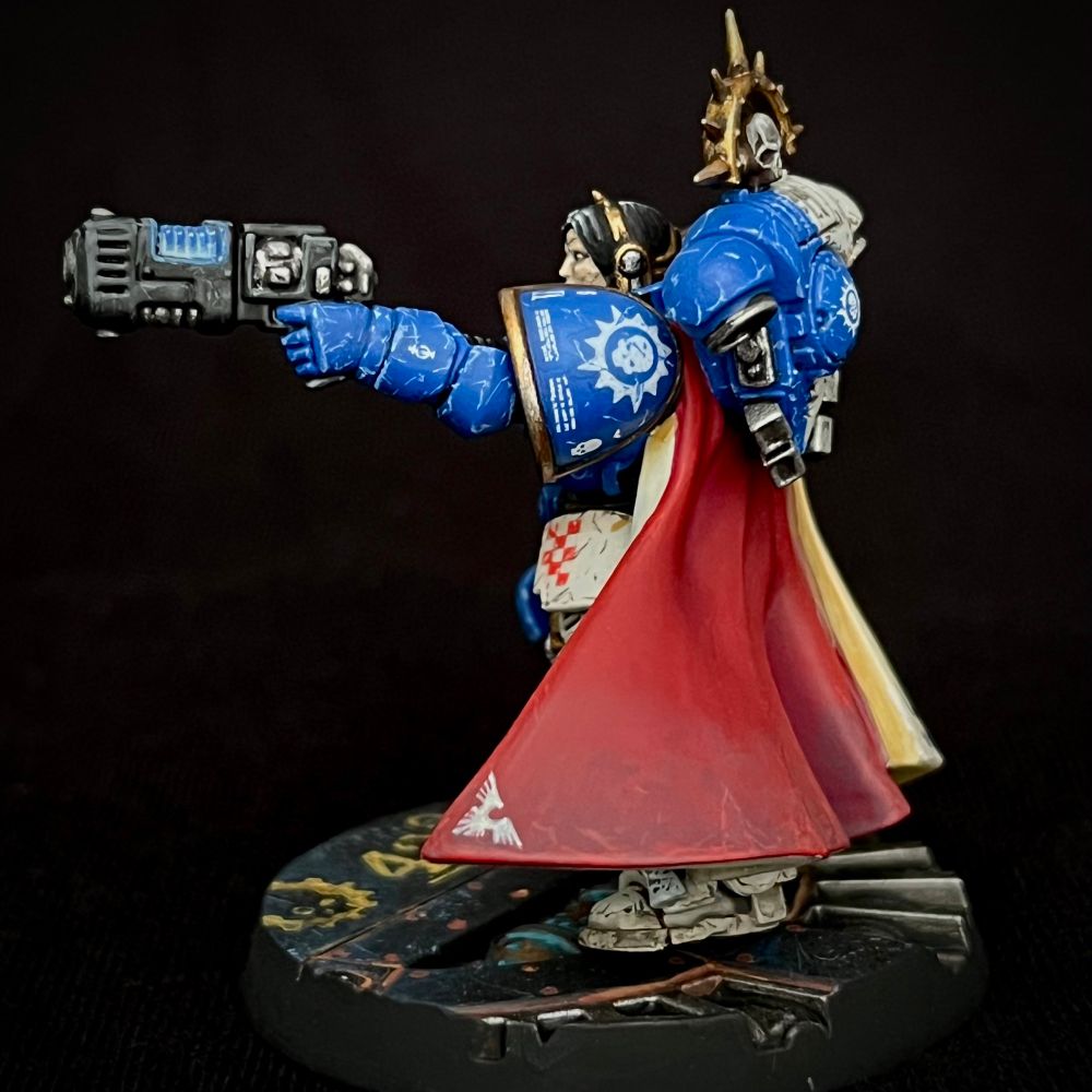A Novamarine Space Marine Captain equipped with a Power Fist and Plasma Pistol. She is wearing quartered bone and blue power armour, a red and cream cloak and is adorned with a lot of decoration and heraldry. She has black hair with a white stripe falling over one eye. All her equipment is battered and worn.