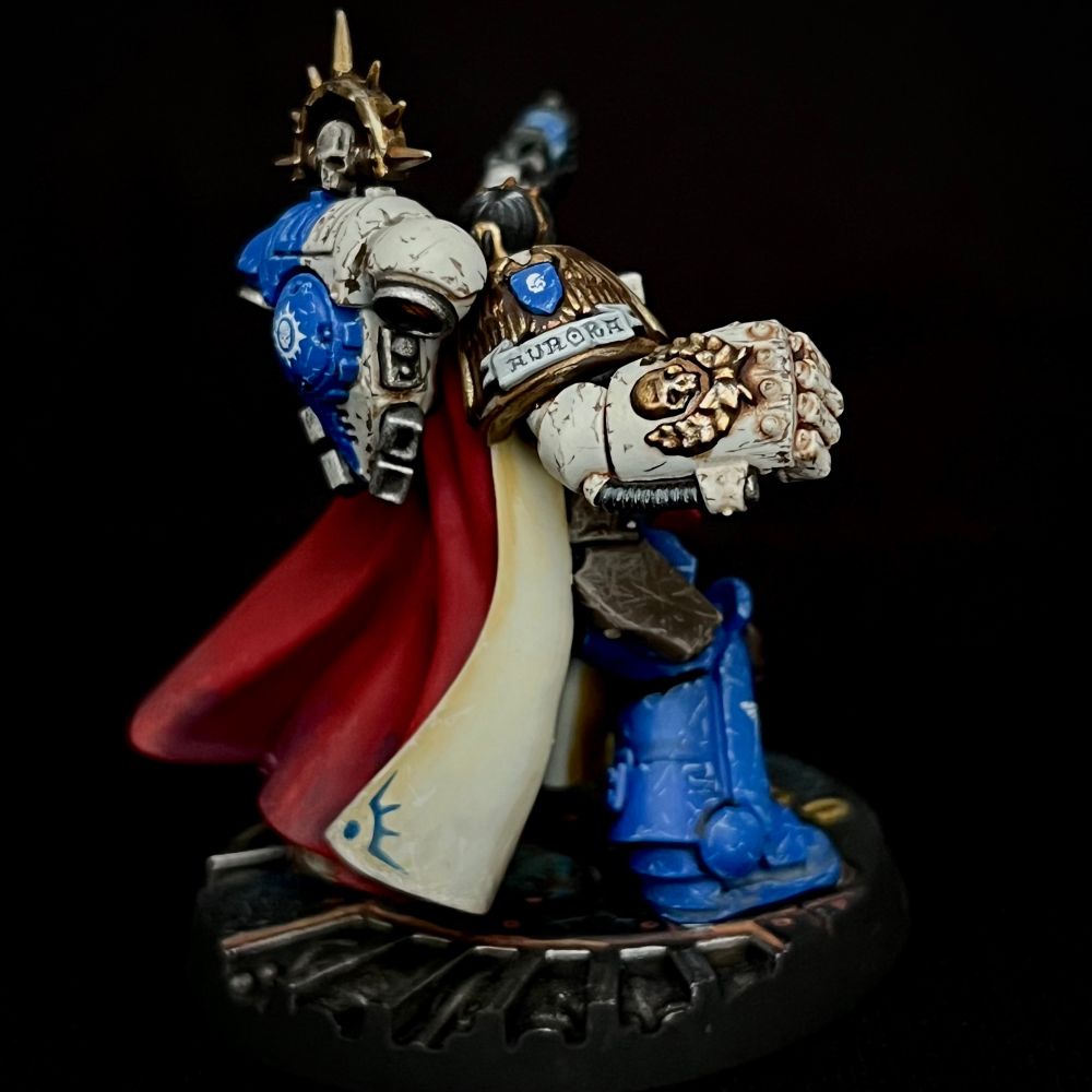A Novamarine Space Marine Captain equipped with a Power Fist and Plasma Pistol. She is wearing quartered bone and blue power armour, a red and cream cloak and is adorned with a lot of decoration and heraldry. She has black hair with a white stripe falling over one eye. All her equipment is battered and worn.