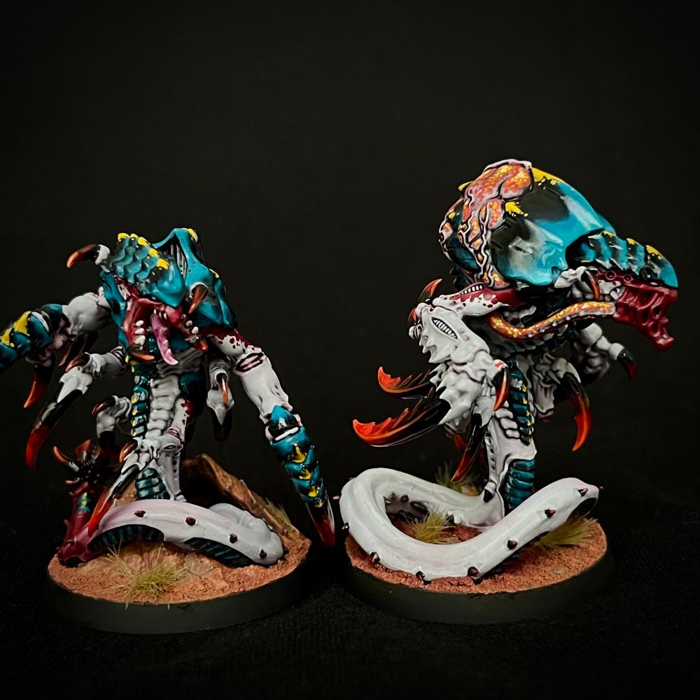 A pair of Hyper-Adapted Raveners painted with glossy black/blue carapace with yellow markings, white/purple skin with red markings, and dark red claws and spikes. One has a split lower jaw, heavier armour and crab-like upper claws, while the other has distended acid-filled sacs on its back, which it can spray from its face
